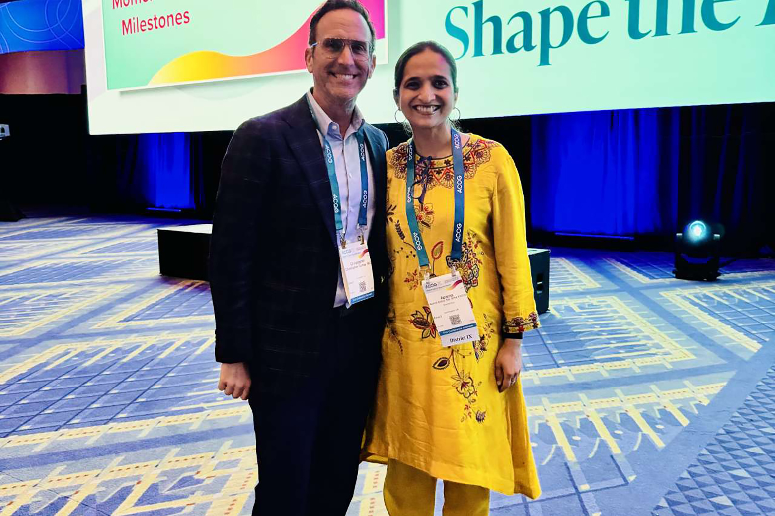 Christopher Tarnay, left, in dark suit and purple button-down, stands next to Aparna Sridhar, in yellow outfit with embroidered details