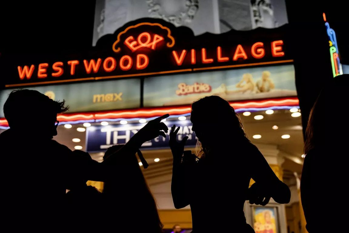 Silhouetted moviegoers use American Sign Language while gathered outside the Regency Westwood Village Theater at night before a special open-caption screening of the ASL version of Barbie in 2023. The theater’s neon marquee glows above the crowd.