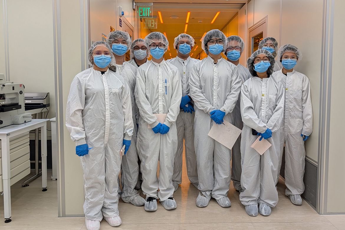 Group of 10 people in cleanroom suits, masks, gloves, and hairnets standing in a laboratory corridor, posing together.