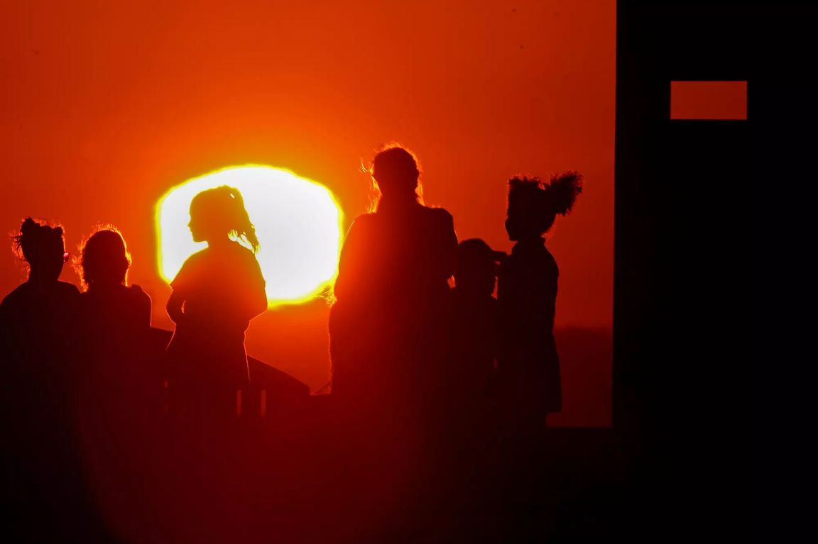 Silhouetted visitors stand atop Signal Hill at sunset as the blazing sun glows orange-red on the horizon, casting the scene in intense light during a heat wave across Southern California in 2022.  