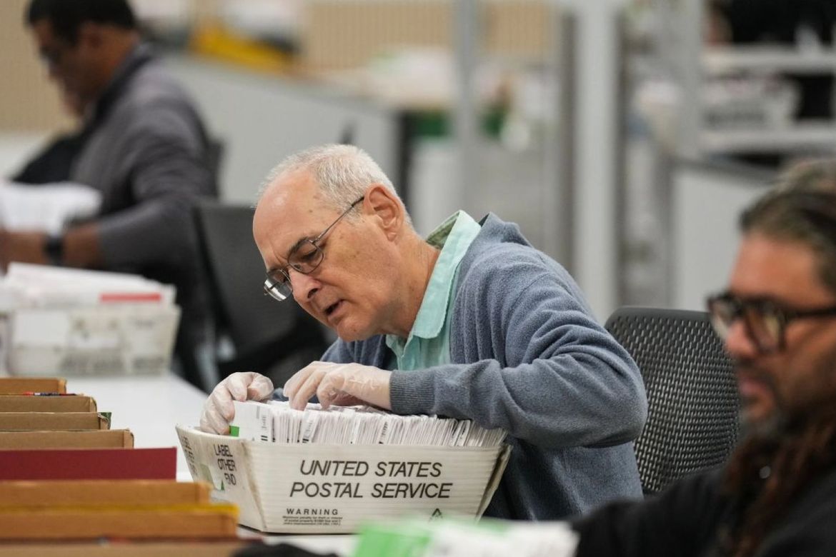 An older male worker in plastic gloves sort ballots in a white USPS mail tray