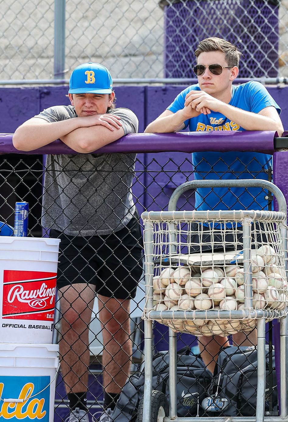 JJ Svenson, right, and a UCLA teammate lean against a purple chain-link fence railing watching practice at the College World Series in Omaha. Svenson wears a blue UCLA Bruins t-shirt and aviator sunglasses. A wire ball cart filled with baseballs and a Rawlings Official Baseball bucket are visible in the foreground.