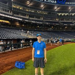 JJ Svenson stands on the field at Charles Schwab Field Omaha during the NCAA College World Series, smiling at the camera. He wears a UCLA Bruins blue polo, blue and gold "B" cap, black shorts, white socks, and blue sneakers, with a credential lanyard around his neck. 