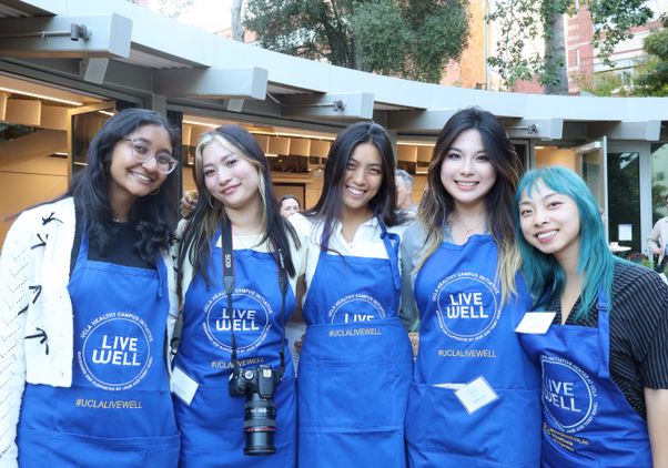 Five UCLA students wearing blue “Live Well” aprons stand together and smile for a group photo at an outdoor campus event. One student has a camera hanging from her neck, and the group poses in front of a courtyard gathering space surrounded by trees and campus buildings.