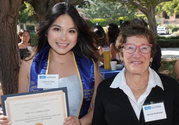 Lumina Chan, wearing a blue Class of 2026 stole, stands beside Marcie Rothman at an outdoor Food Studies celebration. Chan holds a certificate recognizing her as the recipient of the 2025–26 Marcie Rothman Centennial Scholars Undergraduate Scholarship while both smile for the camera beneath shaded campus trees.