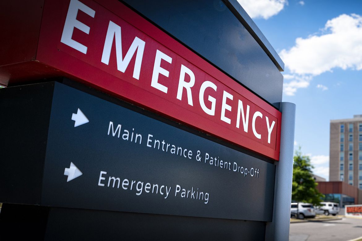 Large red and black hospital sign reading “EMERGENCY” with directional arrows for main entrance, patient drop-off, and emergency parking, blue sky background.