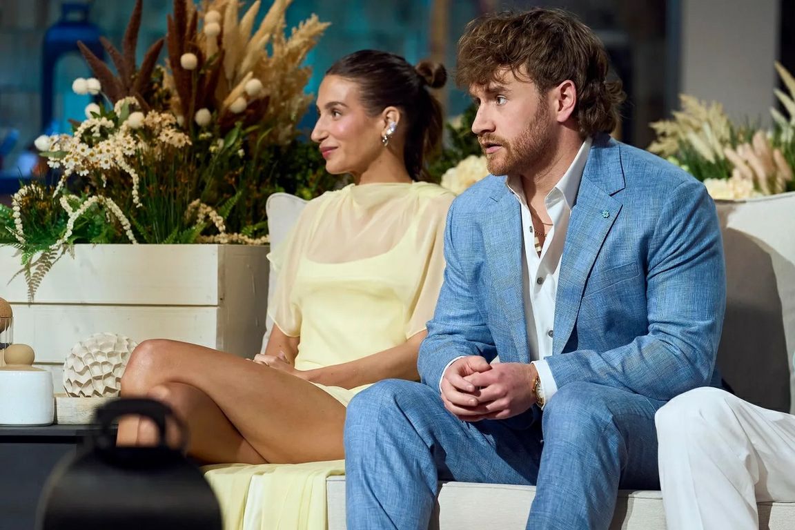 Reality television personality Amanda Batula sits on a sofa during a reunion-style panel discussion, wearing a pale yellow dress and smiling as she listens. Floral arrangements decorate the set behind her, and another cast member sits nearby.