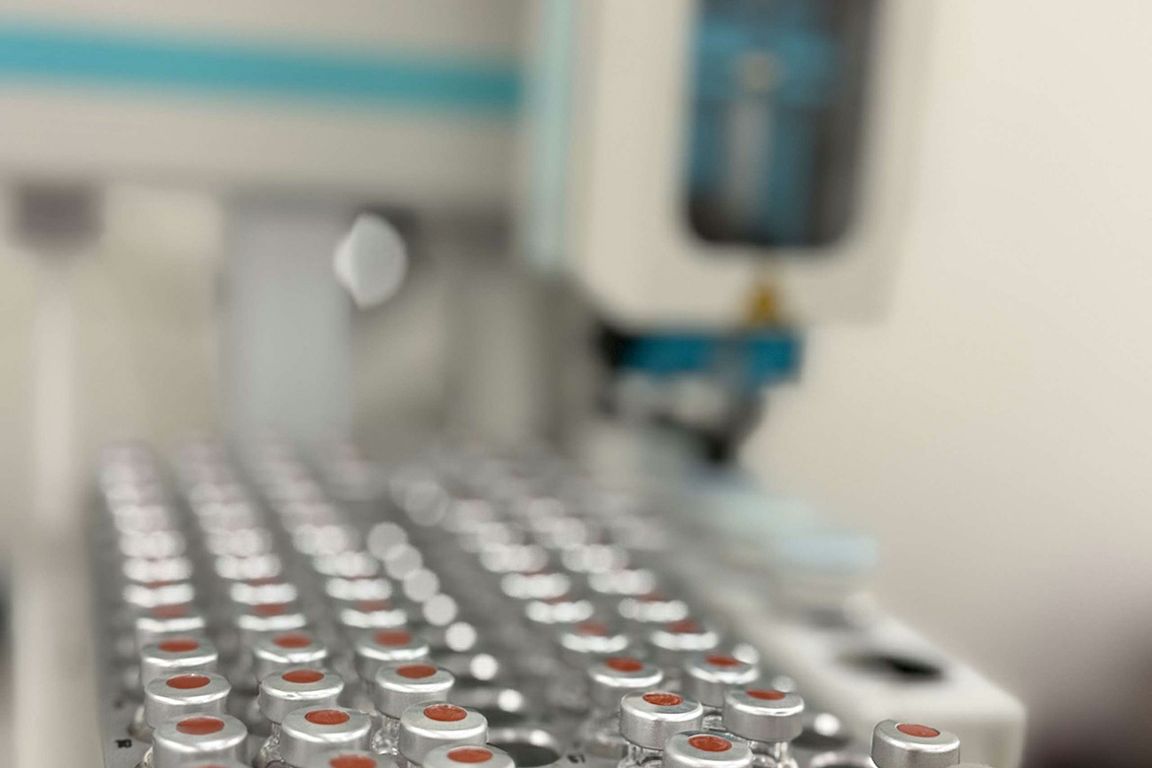 Rows of silver-capped laboratory vials are arranged in a tray, with automated laboratory equipment visible in the background.