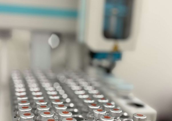 Rows of silver-capped laboratory vials are arranged in a tray, with automated laboratory equipment visible in the background.