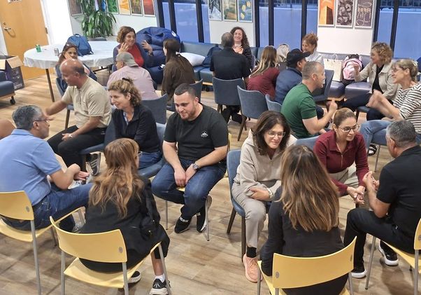 About 20 adults sit in pairs and small groups in a bright community room, facing each other in conversation. Chairs are arranged in circles, creating an interactive workshop atmosphere.