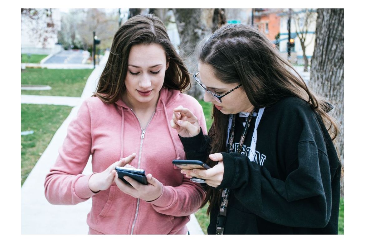 A young women in a pink hoodie touches the screen on her smartphones while second woman in black sweatshirt and glasses looks at the screen