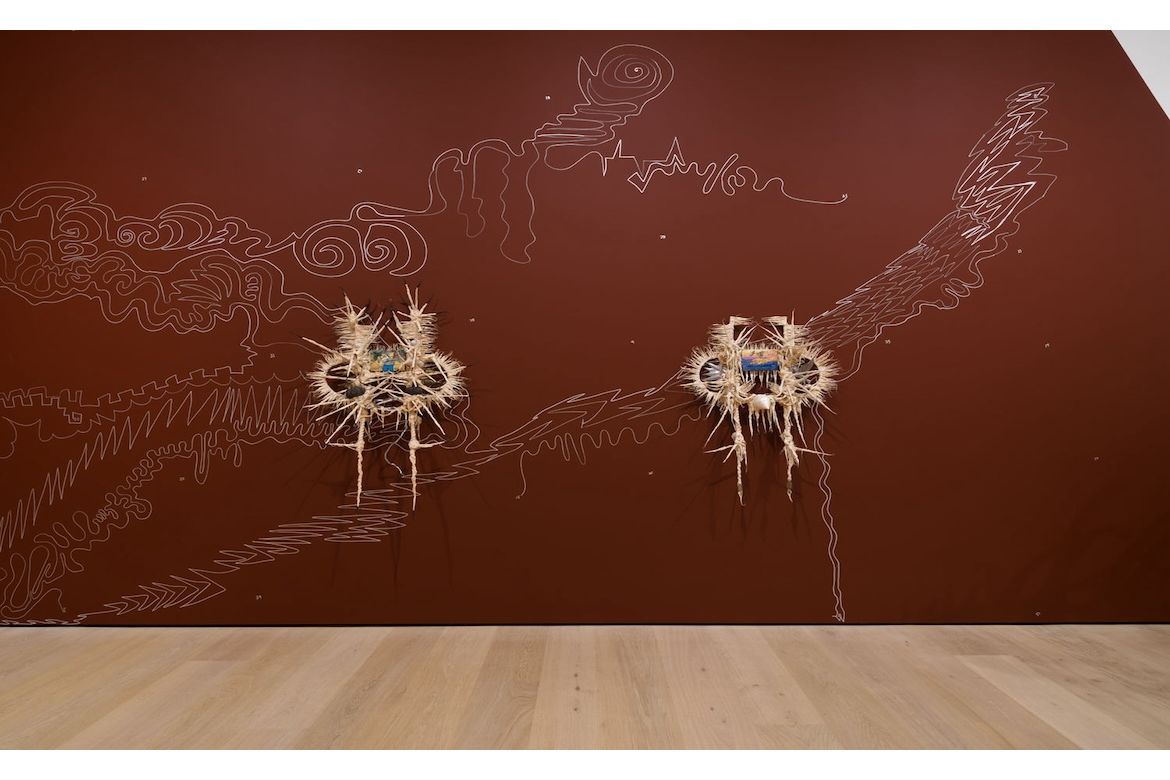 Wall installation with two straw-and-fiber sculptural forms mounted on a reddish-brown wall, connected by white, hand-drawn abstract lines resembling swirling landscapes, waves and lightning across the surface.