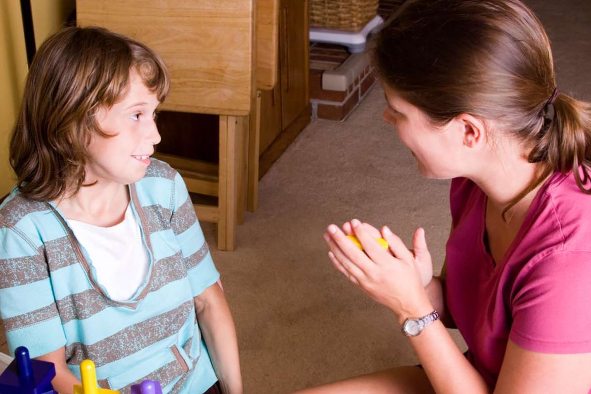 Woman kneeling faces a smiling child indoors, gesturing with clasped hands while speaking. The child sits beside colorful toy blocks in a home setting.