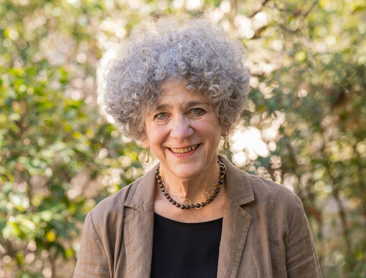 Portrait of UCLA professor emerita Johanna Drucker smiling outdoors, wearing a tan blazer over a black top, with curly gray hair framed by a softly blurred backdrop of trees and greenery.
