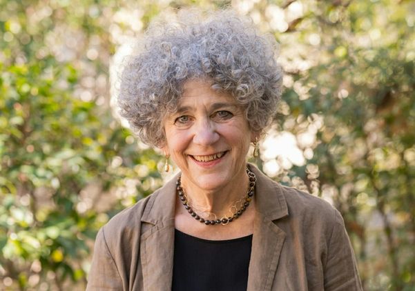Portrait of UCLA professor emerita Johanna Drucker smiling outdoors, wearing a tan blazer over a black top, with curly gray hair framed by a softly blurred backdrop of trees and greenery.