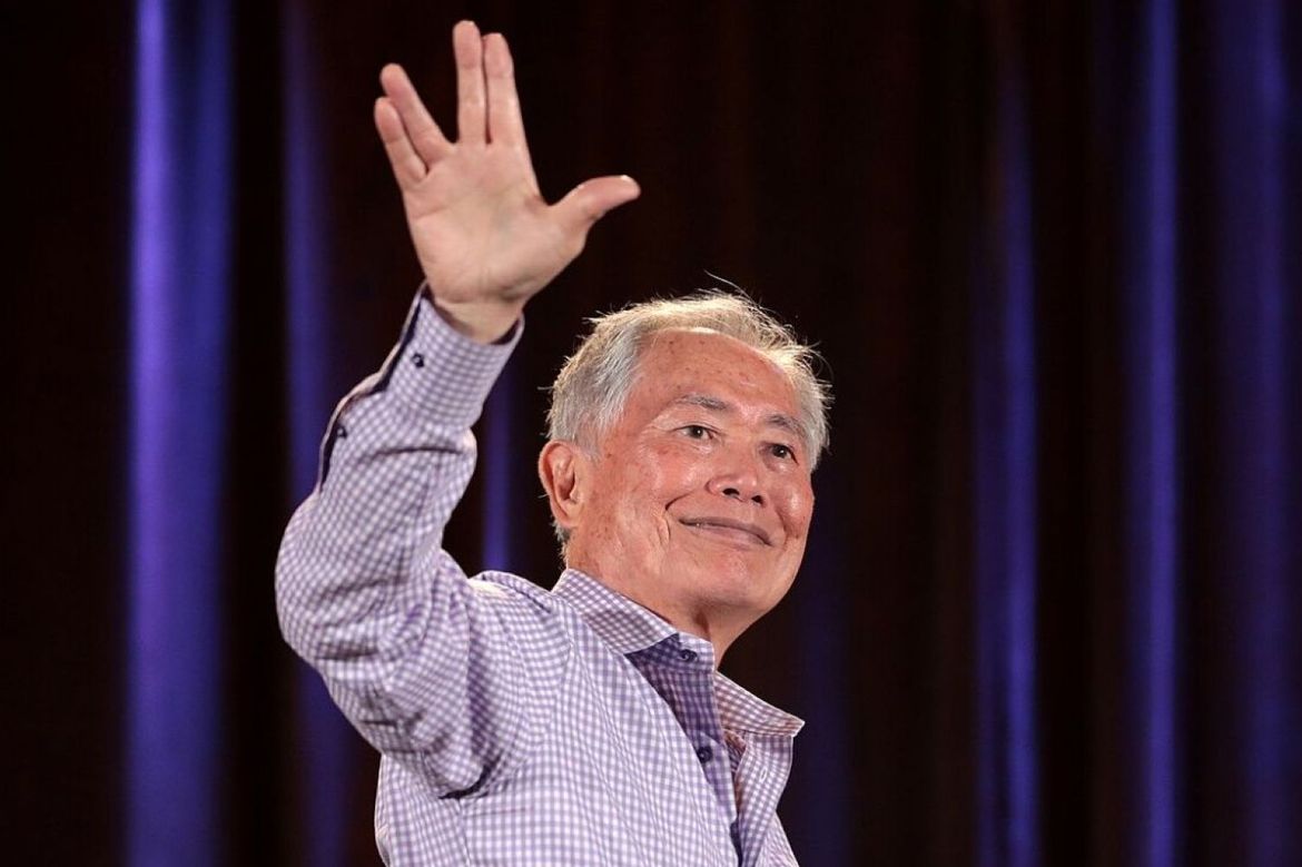 George Takei holds up right hand in signature Star Trek split-finger salute in front of purple stage curtain