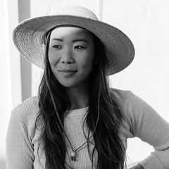 Black-and-white portrait of Hana Asano wearing a wide-brimmed hat and light sweater, with long hair, looking slightly to the side while standing indoors with one hand on her hip.