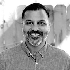 Black-and-white headshot of Alex Bhattacharji, with short dark hair and a trimmed beard, wearing a light button-up shirt, standing in front of a softly blurred wooden fence.