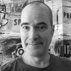 Black-and-white headshot of Dave Plunkert wearing a dark T-shirt in front of a blurred wall covered with posters and graphics.