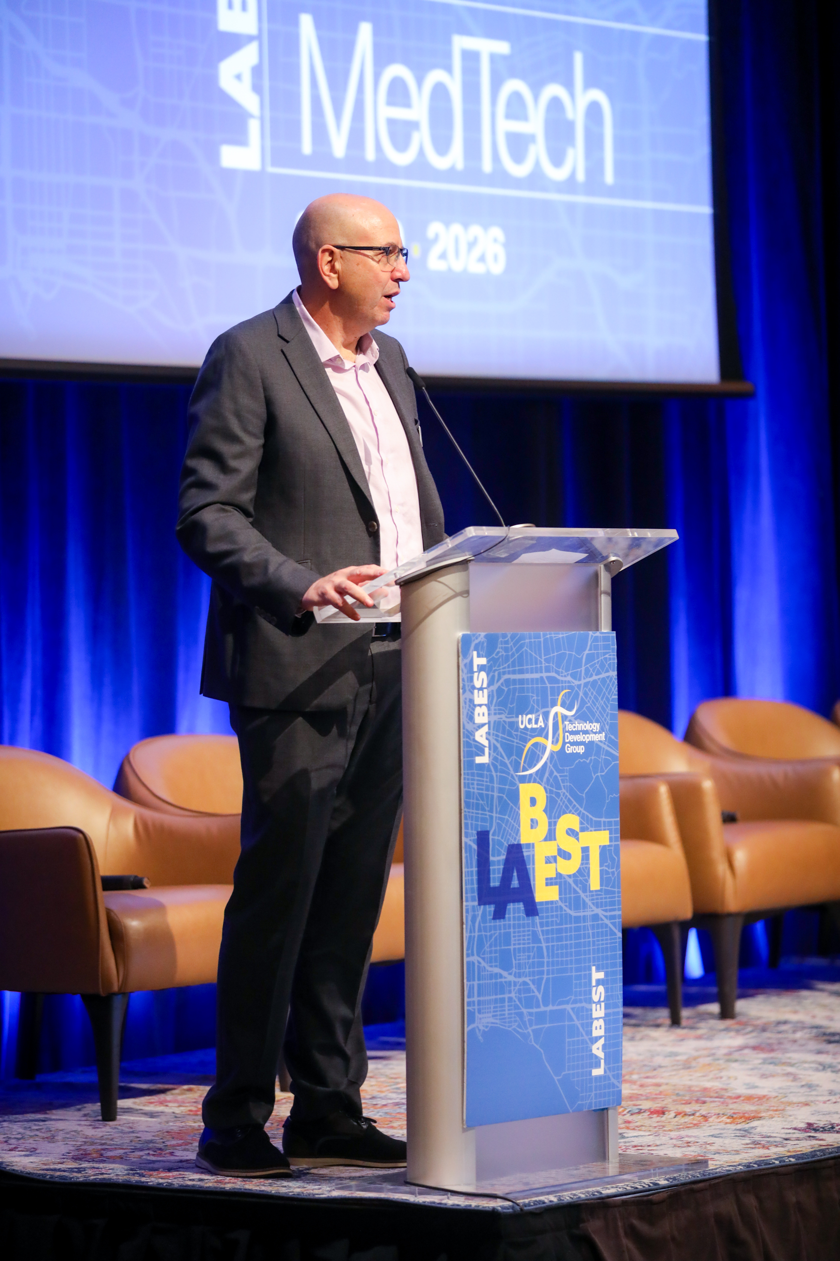 Amir Naiberg stands at a UCLA Technology Development Group podium during the 2026 LABEST MedTech conference, addressing attendees on a stage framed by blue lighting. A large screen behind him displays conference branding, while panel chairs sit nearby.