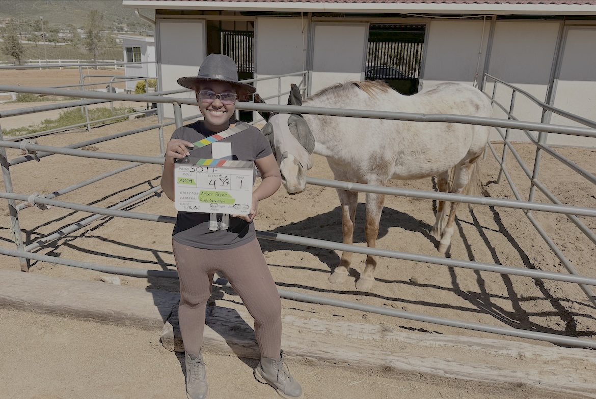 Amber Payne stands beside a horse enclosure holding a film clapperboard, smiling toward the camera, wearing hat and casual clothing.