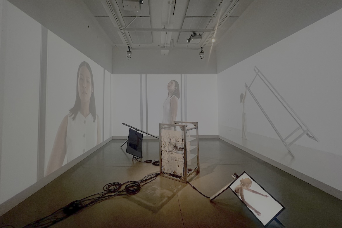 A white room with computer hardware and screens, surrounded by video screens featuring a woman in white.