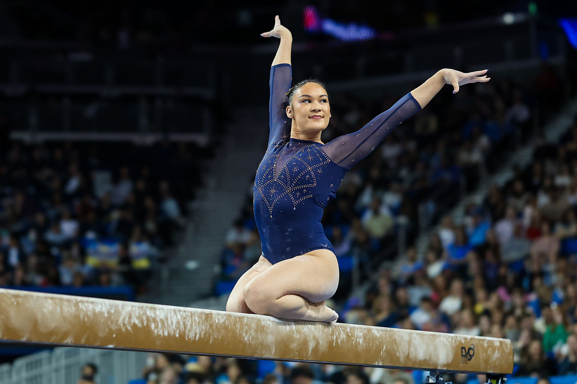 UCLA gymnast Ciena Alipio performs on the balance beam, seated with one leg tucked, arms raised overhead and extended to the side, wearing a navy blue long-sleeve leotard with rhinestone detailing, before a packed arena crowd.