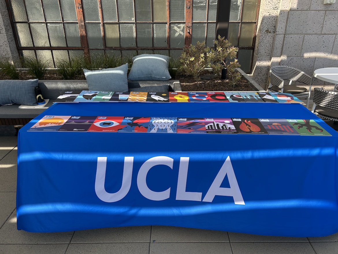 A table draped with a UCLA-blue cloth displays copies of Blueprint magazine on the rooftop of UCLA Downtown. Two rows of colorful magazine covers are arranged for guests attending the outdoor civic conversation event.