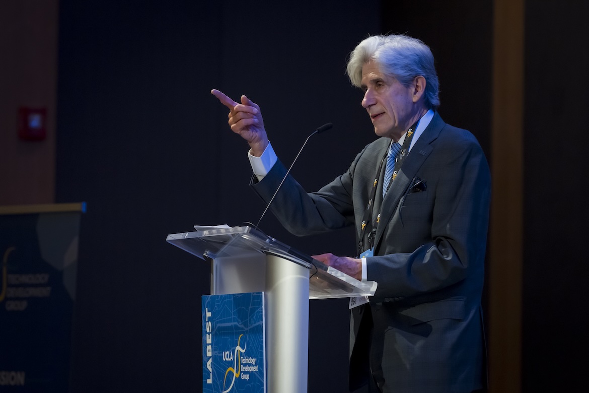 Julio Frenk speaks from a podium during a UCLA Technology Development Group event, gesturing toward the audience while delivering remarks. Stage lighting highlights the chancellor against a dark auditorium backdrop, with UCLA and LABEST branding visible on the lectern.