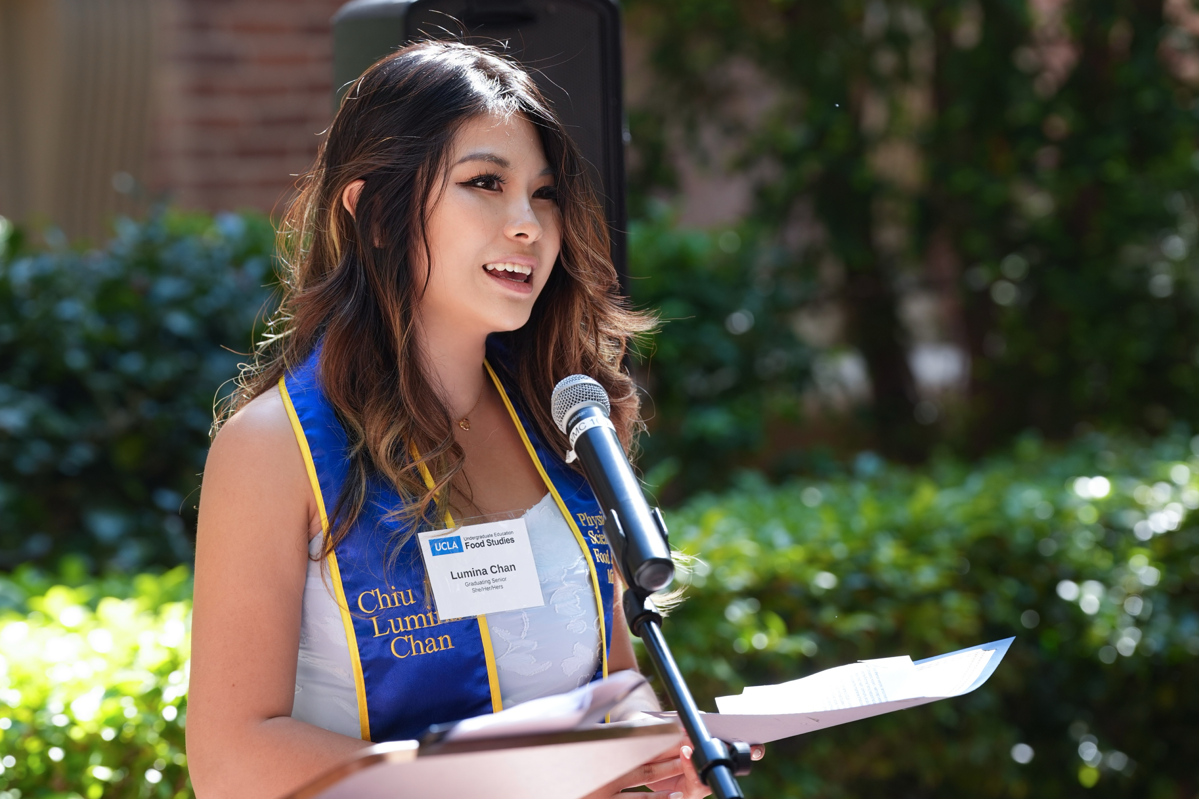 Lumina Chan speaks at an outdoor Food Studies celebration, standing at a microphone and reading from prepared remarks