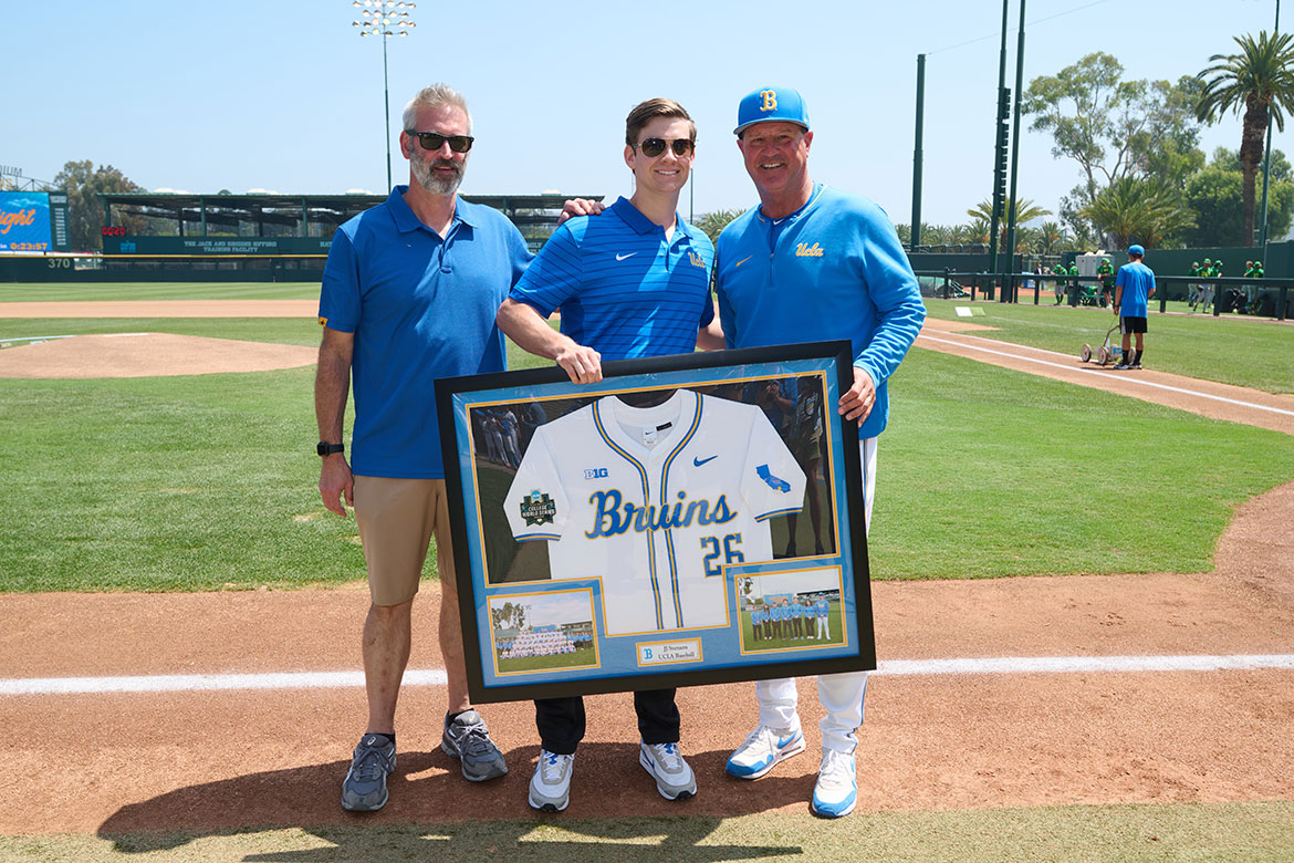 L-R Erik Svenson, JJ Svenson and UCLA baseball head coach John Savage on the baseball diamond, holding a framed jersey