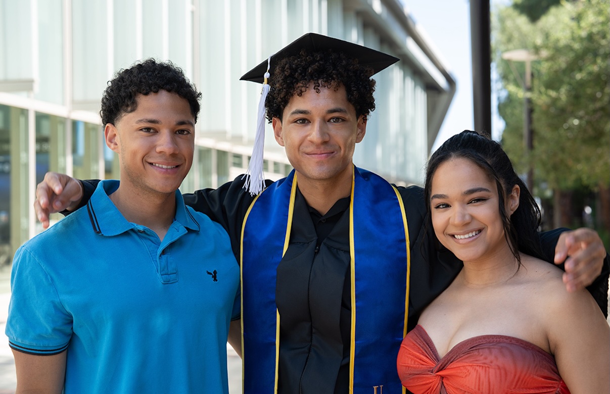 UCLA graduate Justin Dixon, wearing a cap, gown and blue stole, stands smiling between his siblings on campus. With their arms around one another, the three pose for a celebratory graduation portrait outside a campus building.