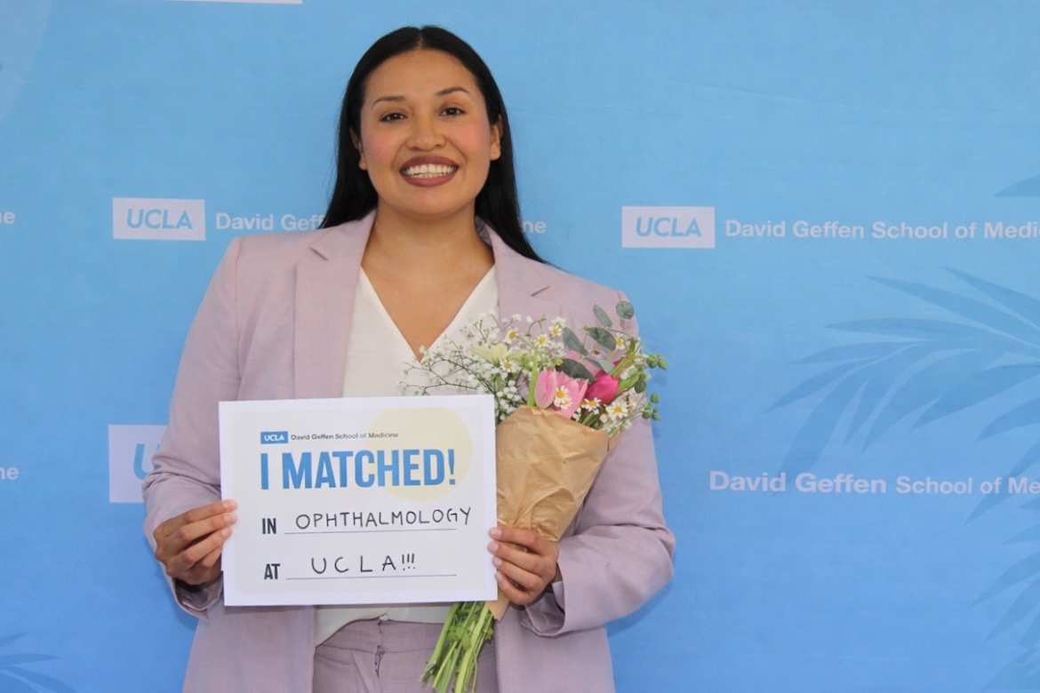 Karla Murillo smiles while holding flowers and a sign announcing her match in ophthalmology at UCLA during Match Day.