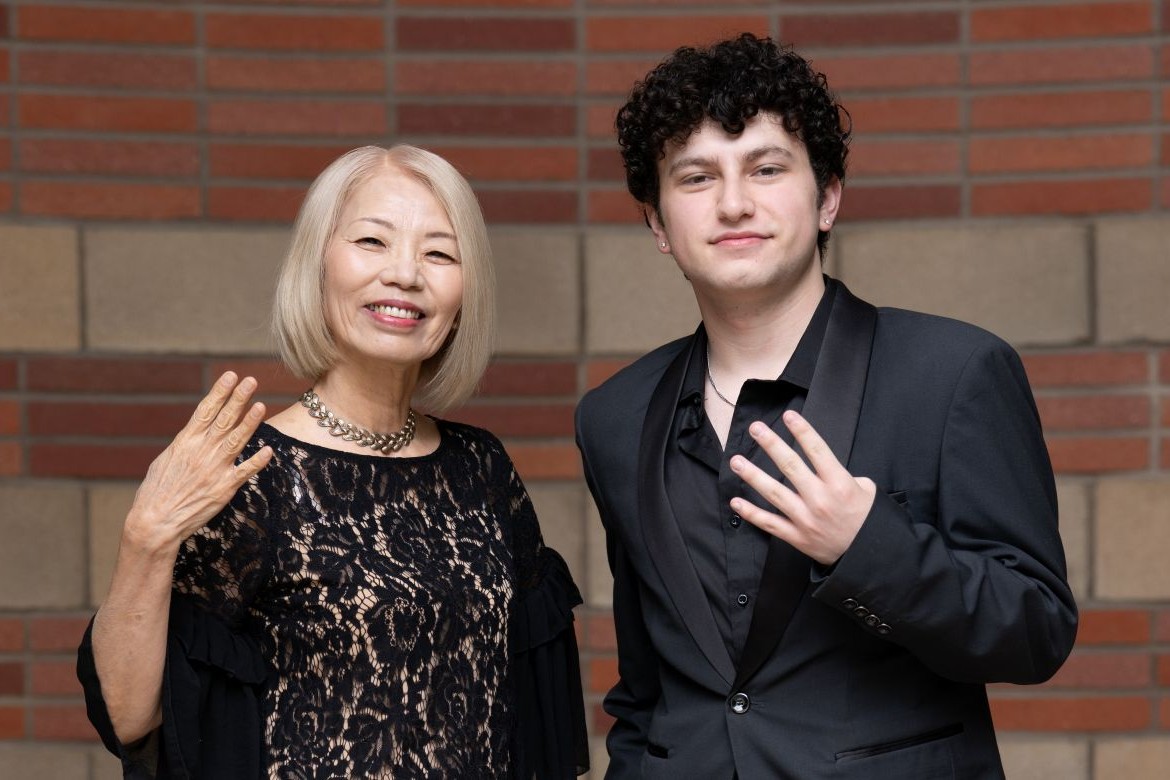 Mayu Sasame and August Maturo stand against brick wall, each smiling and holding up four fingers