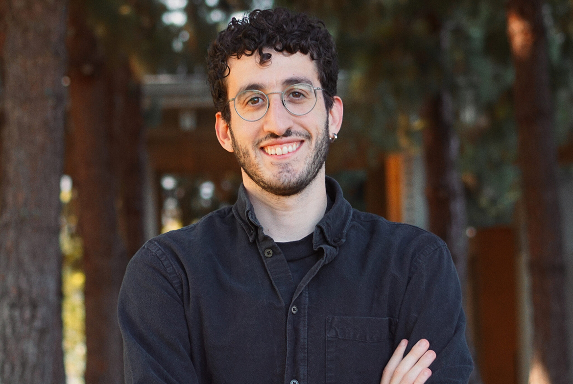 Head-and-shoulders image of Paul Melas with arms crossed while wearing a dark blue button-down and glasses. Trees in the background.