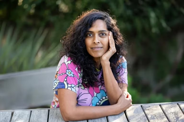 Priya Swaminathan sits at an outdoor wooden table, resting her chin on one hand and looking toward the camera. She wears a colorful floral-patterned shirt