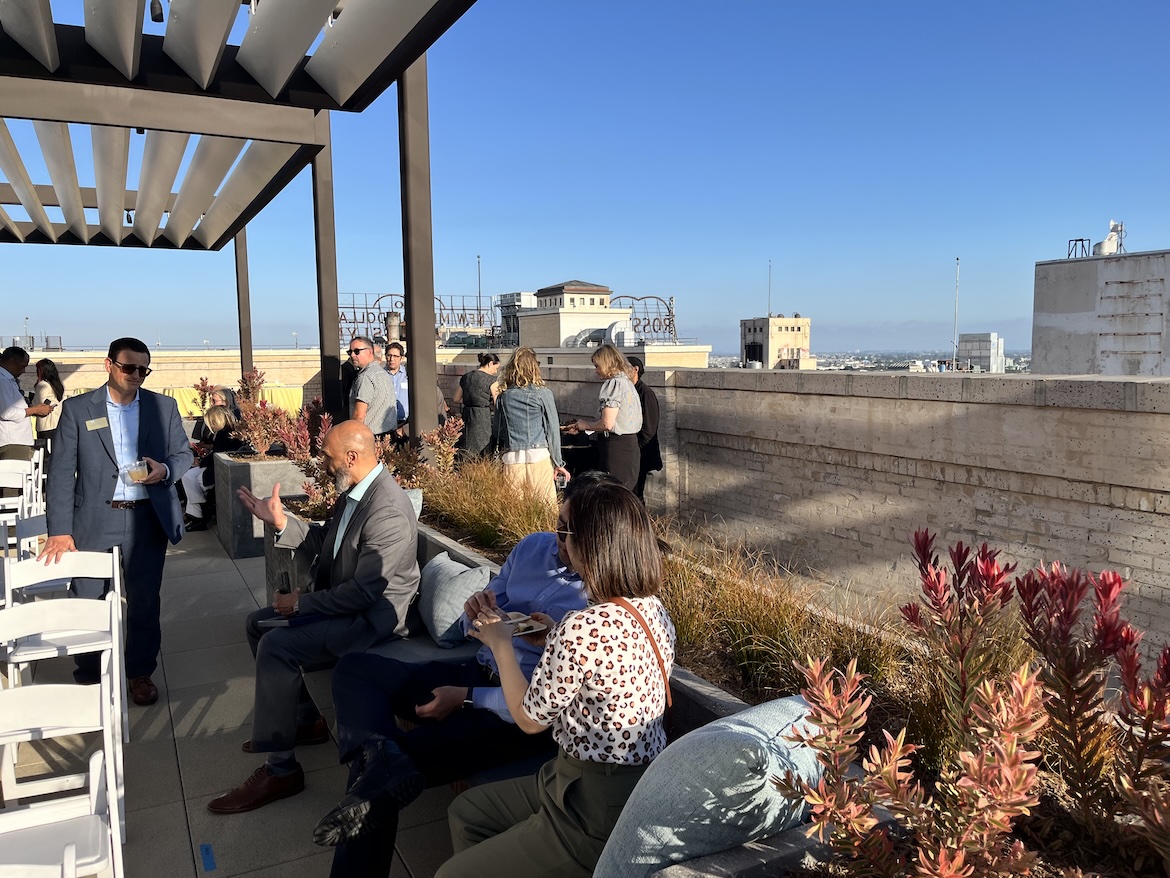 People gather and converse on the rooftop terrace of UCLA Downtown, with seating, planters and pergolas overlooking the Los Angeles skyline under a clear blue sky. The elevated setting provides a panoramic backdrop for the outdoor event.
