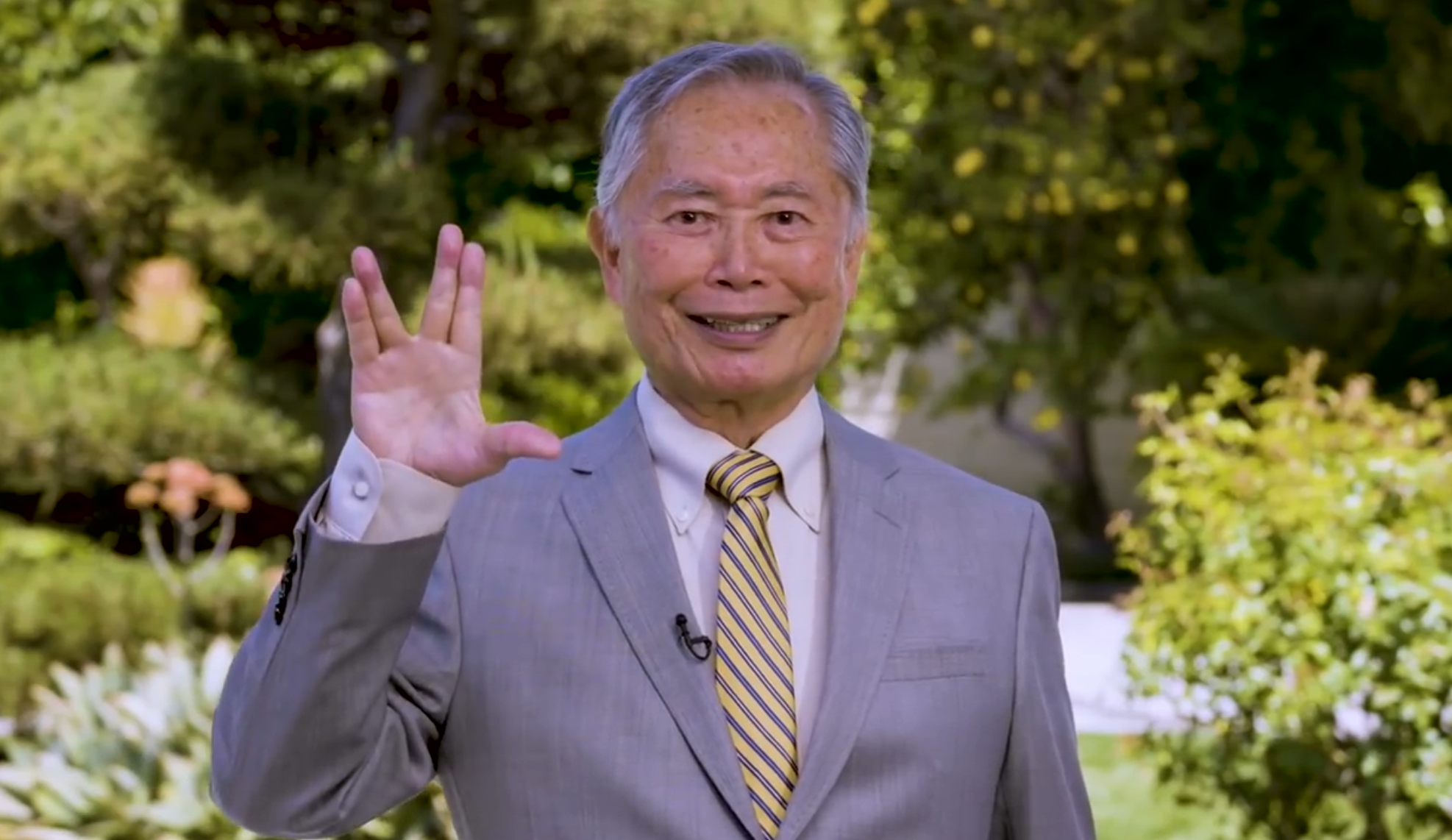 George Takei holds up right hand in Star Trek-style Live Long and Prosper hand signal in front of lush green lawn and bushes 