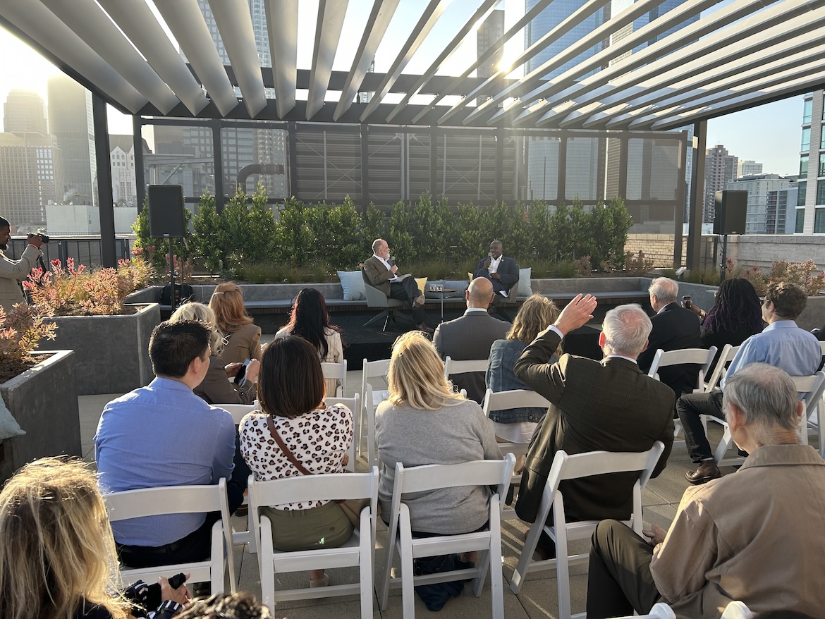Audience members attend a rooftop conversation at UCLA Downtown, where Jim Newton interviews Los Angeles City Council President Marqueece Harris-Dawson before the downtown skyline. Late-afternoon sunlight filters through the pergola, illuminating the outdoor gathering.
