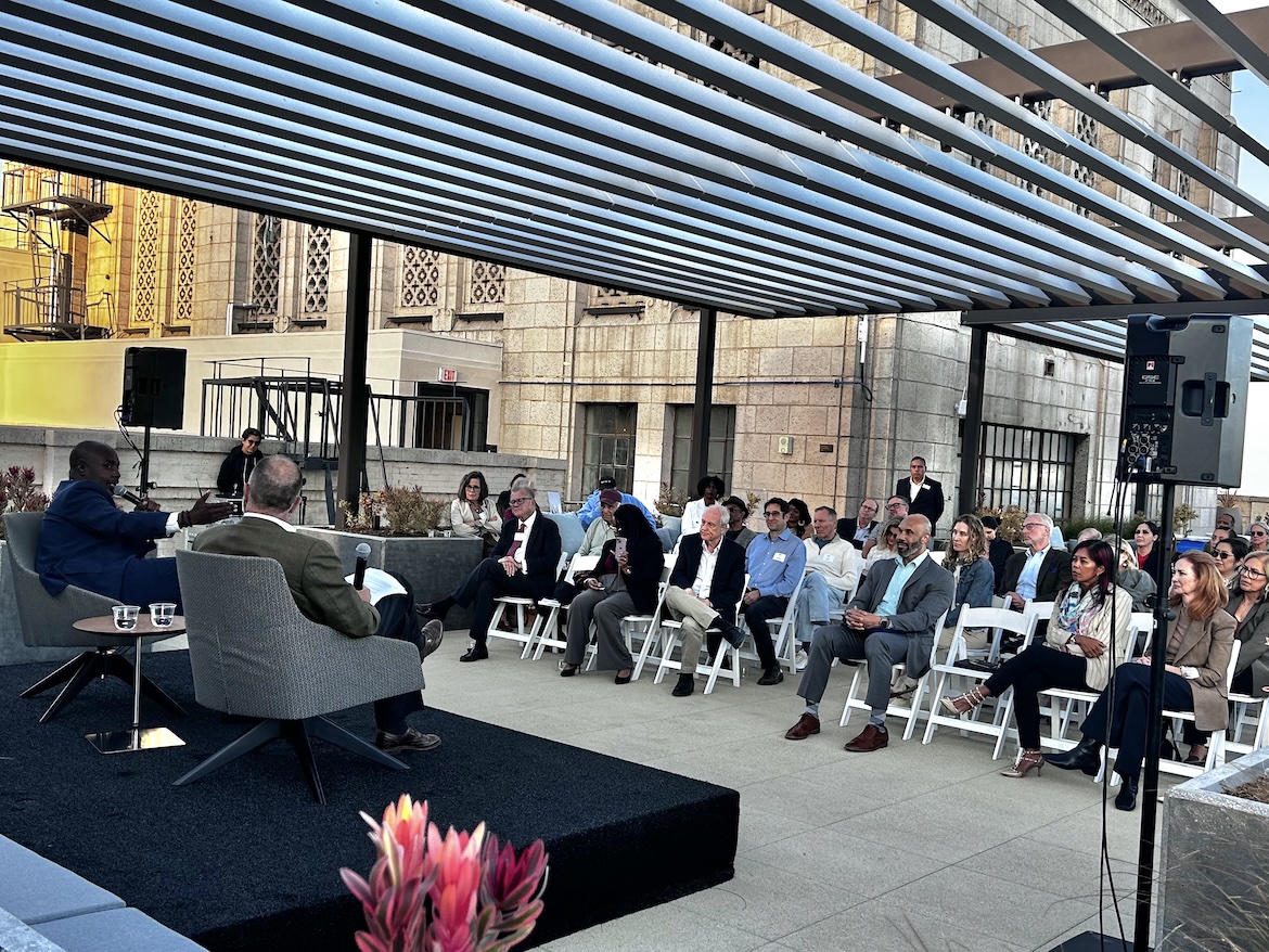 From behind, UCLA's Jim Newton speaks with Los Angeles City Council President Marqueece Harris-Dawson on the rooftop of UCLA Downtown as attendees listen from rows of chairs beneath a pergola, with surrounding downtown buildings framing the outdoor discussion.