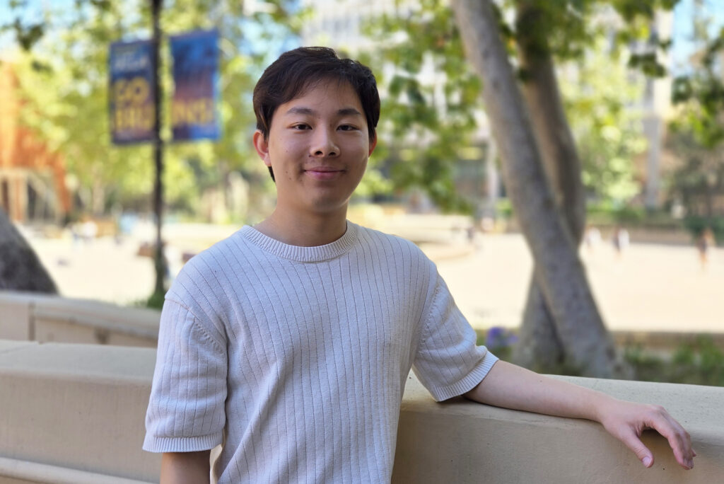 Student Ziyan Xie stands outdoors on the UCLA campus, resting one arm on a low stone wall and smiling at the camera. Trees and campus walkways blur softly in the background.