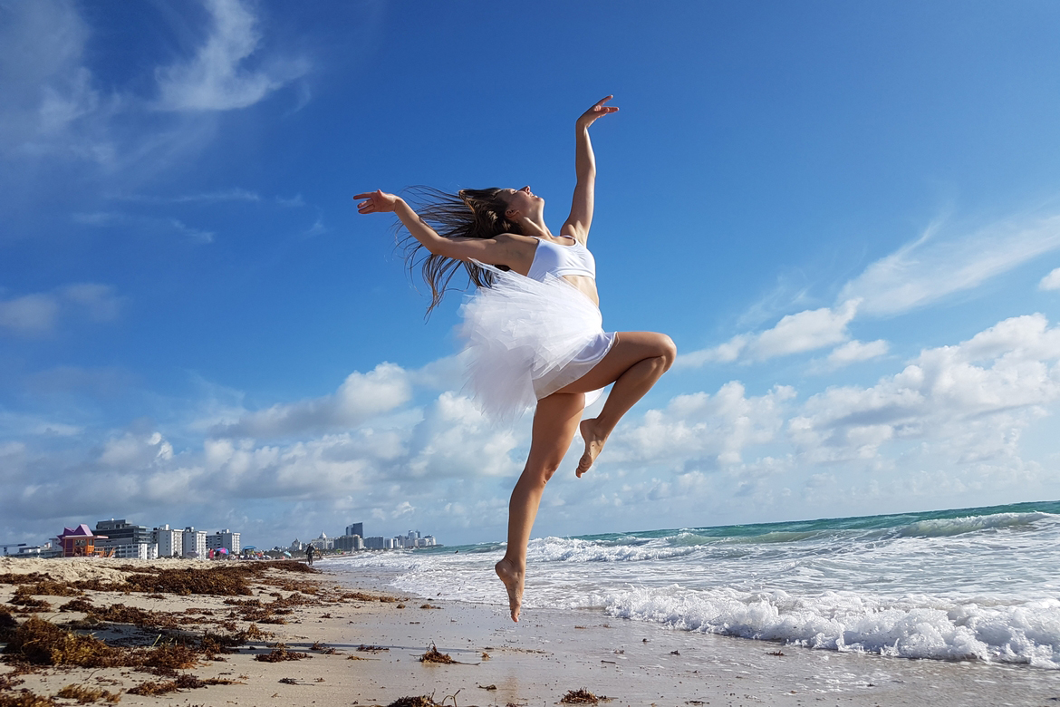 Angel Hass in a white ballet-inspired outfit leaps gracefully above a sandy beach, arms extended and hair flowing. Ocean waves, scattered seaweed, distant buildings, and a bright blue sky with clouds frame the dynamic scene.