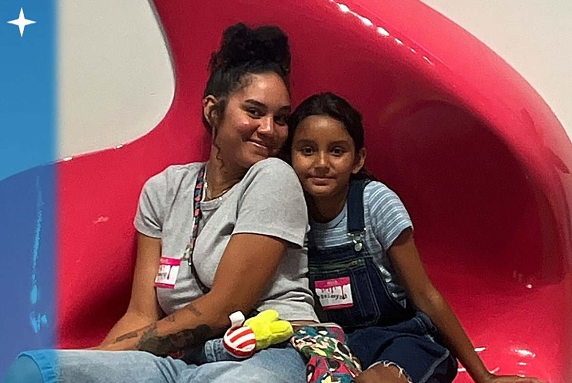 Dejanay Wallace sits with her young daughter on a bright pink sculptural chair, smiling together during a community event. Both wear name tags and sit side by side closely.