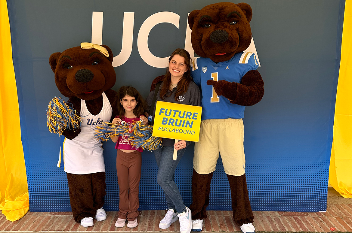Tahlia Disisto and her daughter pose with UCLA mascots Josie and Joe Bruin in front of a UCLA backdrop. Tahlia holds a yellow sign reading “Future Bruin #UCLABOUND” while her daughter holds blue-and-gold pom-poms.