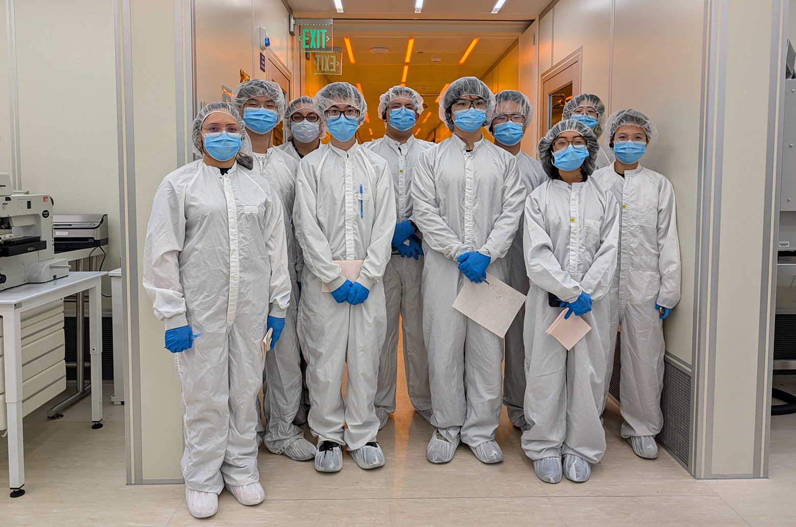 Group of 10 people in cleanroom suits, masks, gloves, and hairnets standing in a laboratory corridor, posing together.