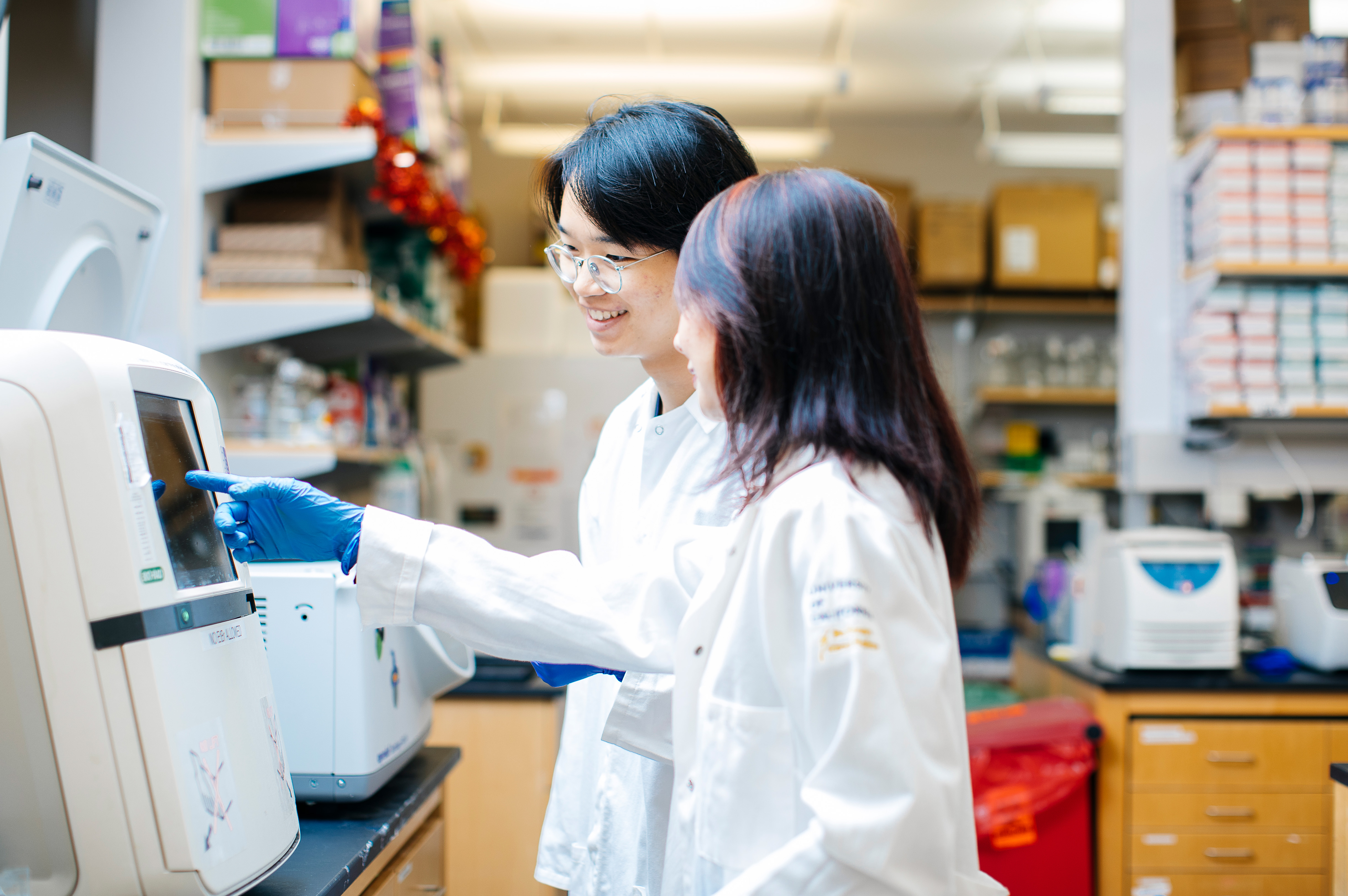 Eliot Kang, with glasses, and Lili Yang wear lab coats and gloves as they review data on equipment in a scientific research lab.