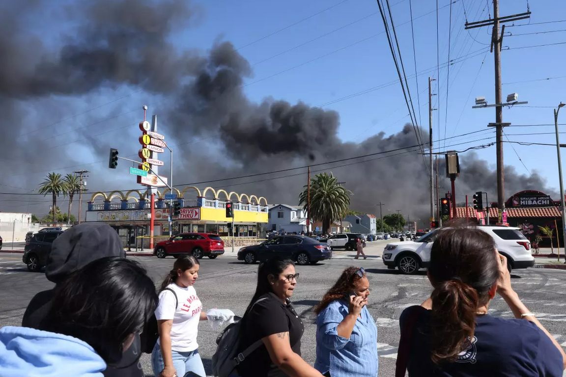People cross South Indiana Avenue in Boyle Heights as thick black smoke billows from a fire at a nearby cold storage facility, filling the sky above the neighborhood.
