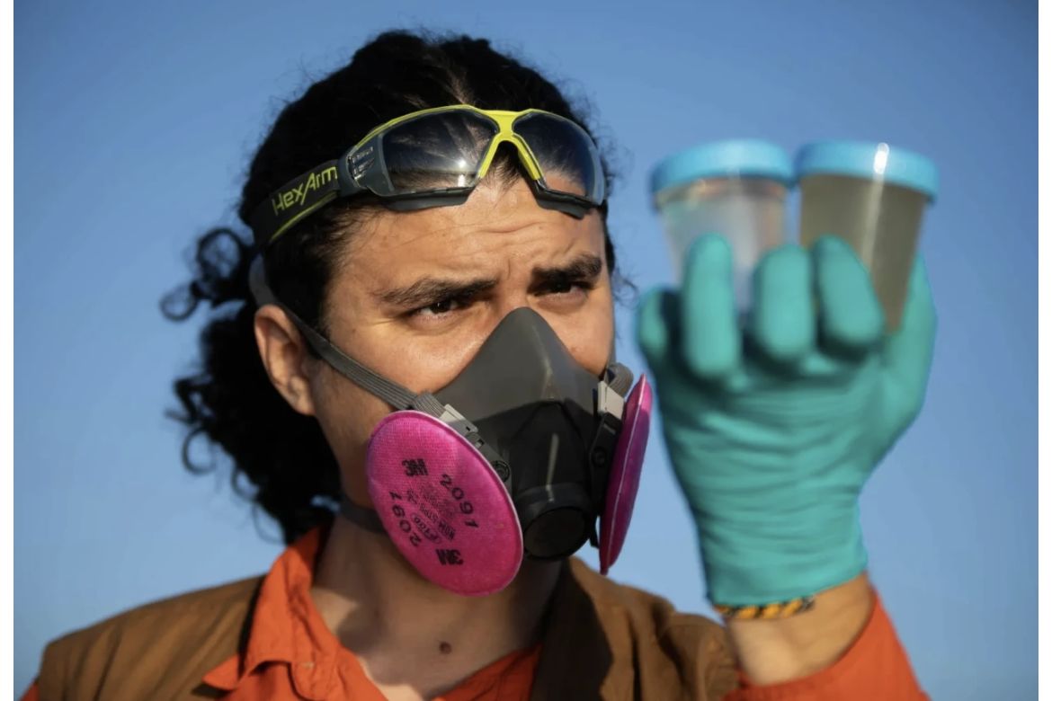 Emmanuel Carrera Ruedas wearing blue plastic gloves and pink facemask holds up two plastic water sample containers with blue lids
