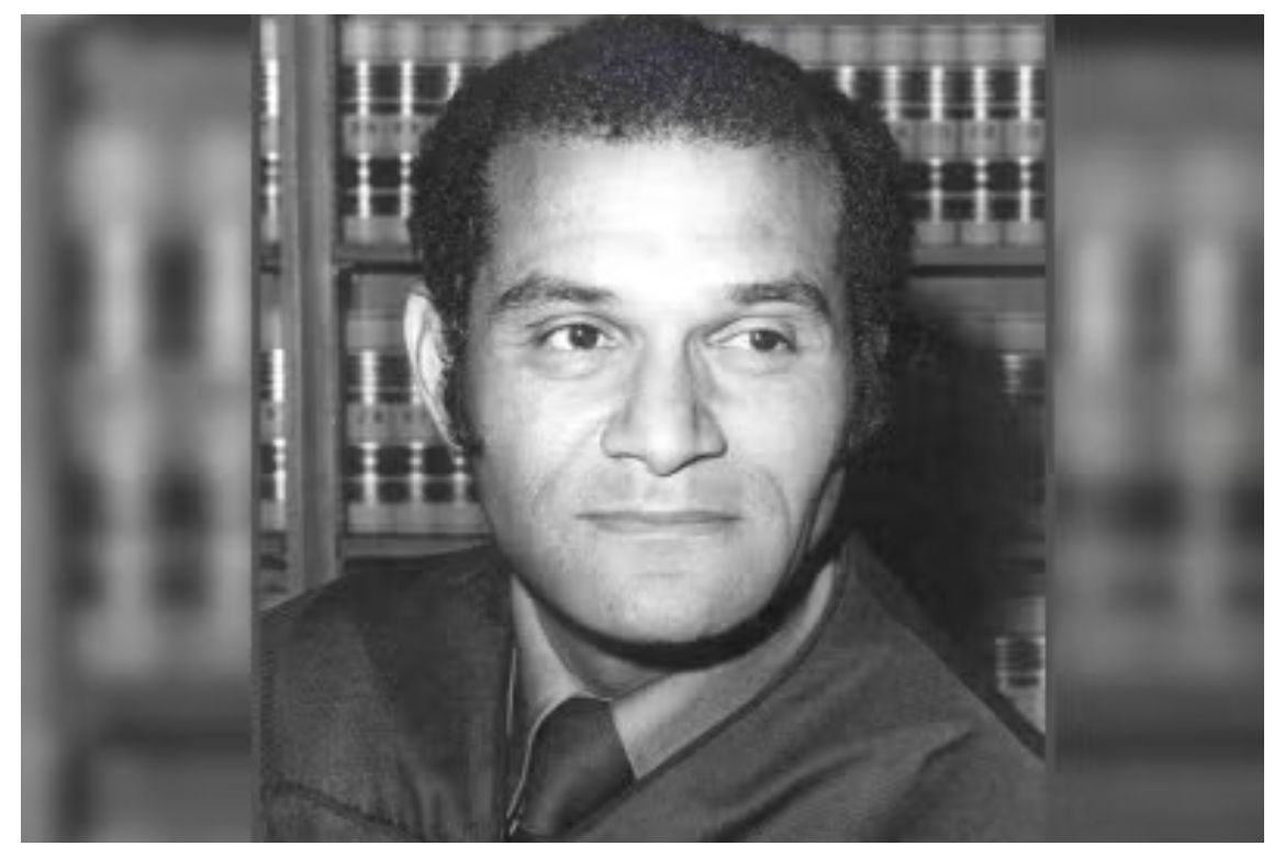 Black-and-white headshot of Billy Mills in black law robe sitting in front of wall of law books