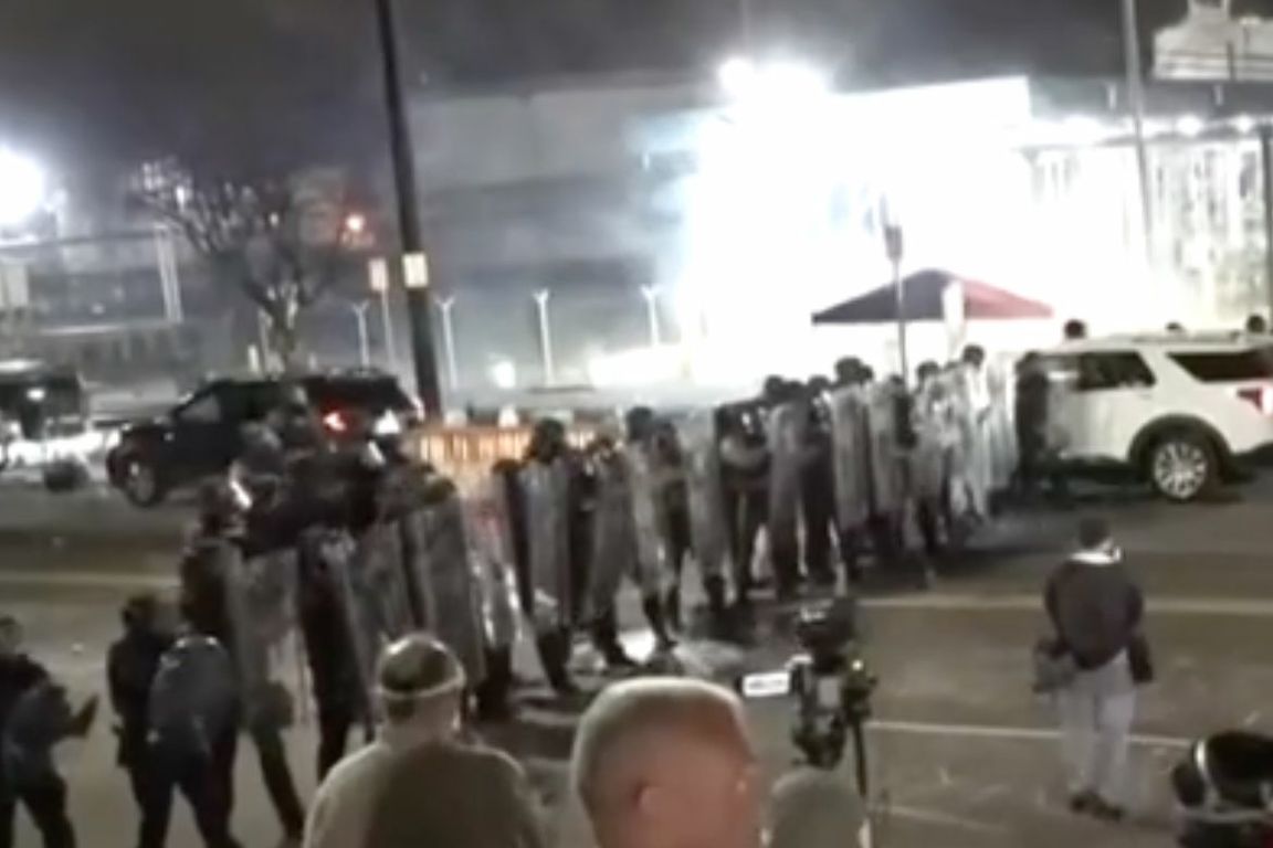 A line of people in protective gear holding shields stands in formation on a street at night, facing a crowd of onlookers.